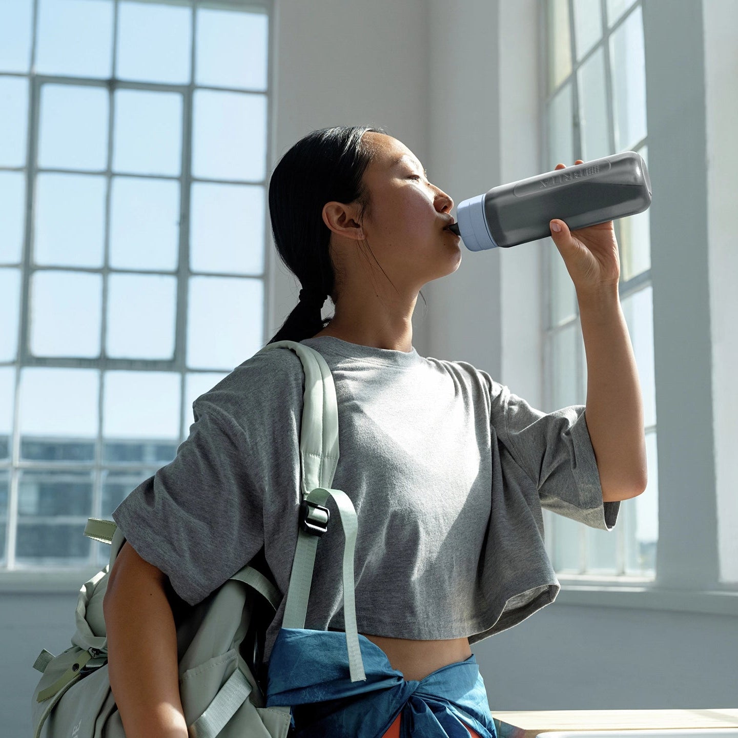 Une femme en train de boire de l'eau d'une carafe BRITA filtre Modèle « Bottle » dans une pièce lumineuse avec de grandes fenêtres.