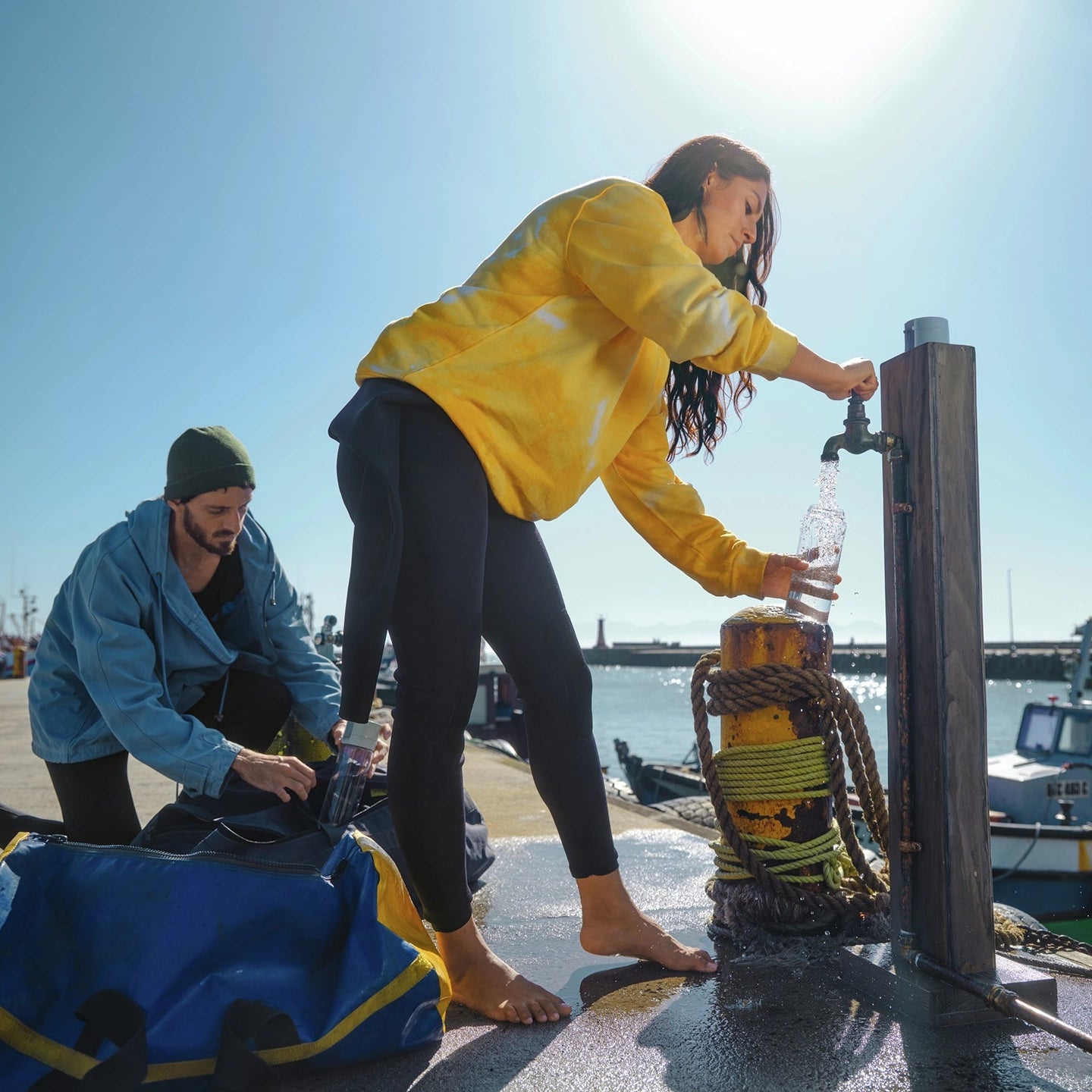 Deux personnes sur un quai au bord d'un plan d'eau ; l'une d'elles utilise un robinet pour remplir une carafe BRITA filtre Bouteille modèle Vital.