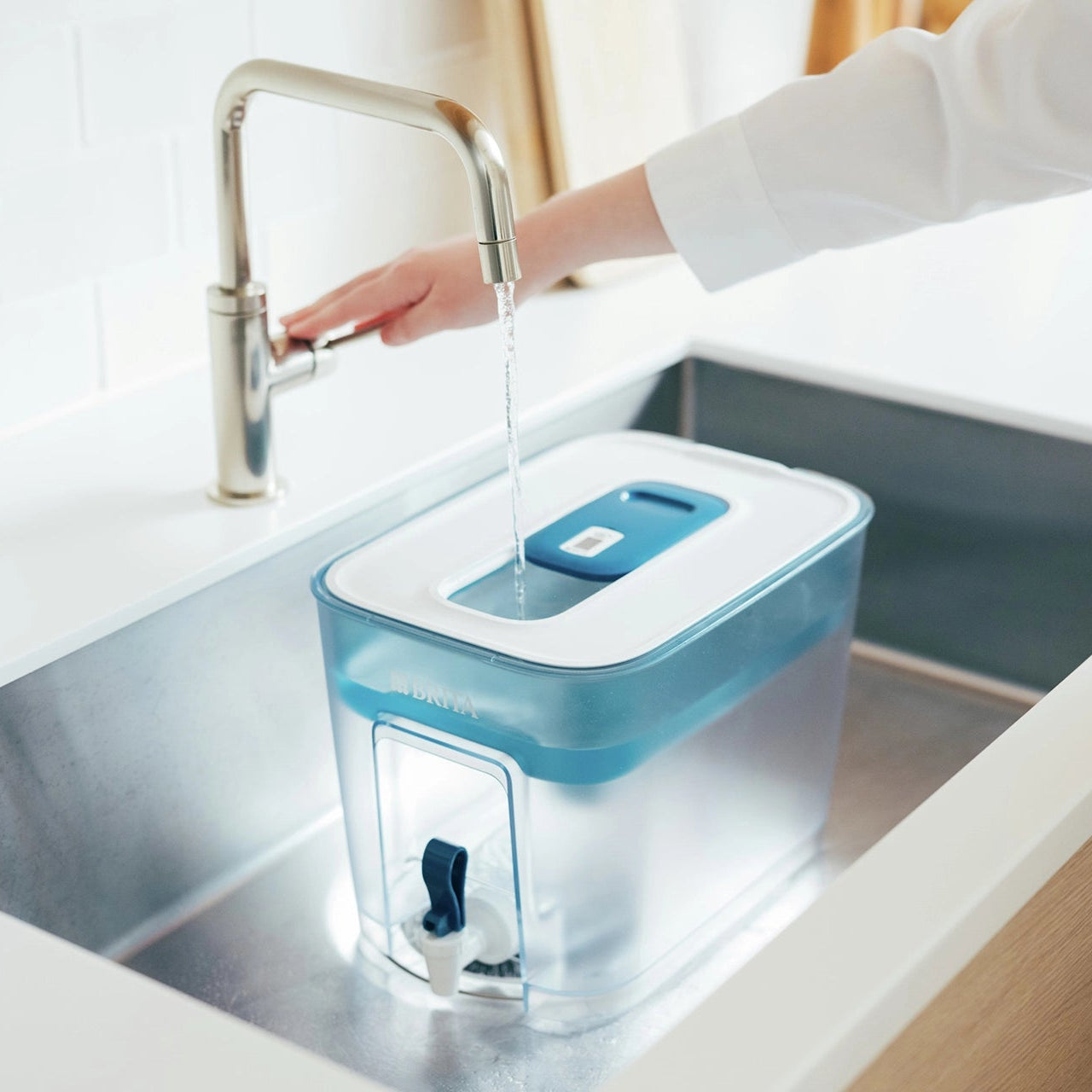 Un filtre à eau BRITA Flow en train d'être rempli d'eau dans un évier de cuisine, sous un robinet ouvert.