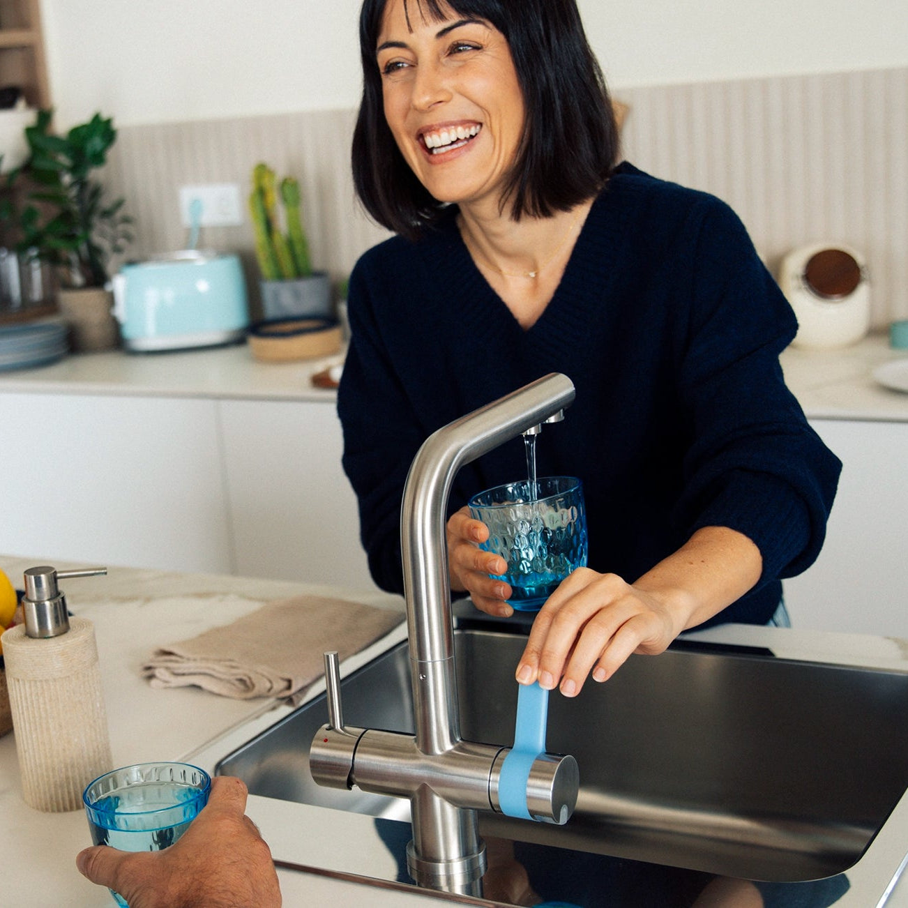 Une femme qui verse de l'eau à partir d'un filtre à eau BRITA à 3 sorties filtre verse un verre dans la cuisine, avec un homme à côté d'elle.